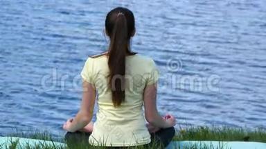 年轻女子在海滩上放松冥想。 坐在草地上打坐在河边做瑜伽的女孩